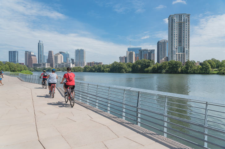 Cyclists ride along a waterfront path with a city skyline in the background, under a blue sky.