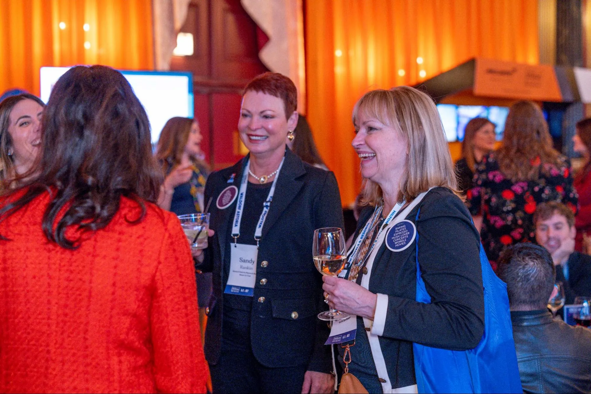  Four female conference attendees talking, smiling, laughing, and holding drinks, during a conference happy hour.